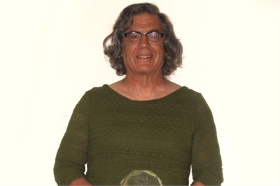 [ai] A person with shoulder-length curly hair, wearing a green top, holding a clear award in front of a plain white background. The individual is smiling and appears to be in a celebratory context.